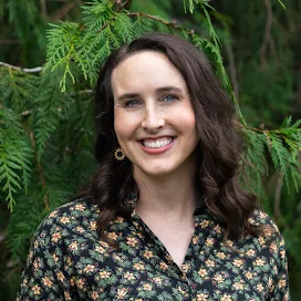 Elsie a femme with dark shoulder length hair smiles brightly against green trees