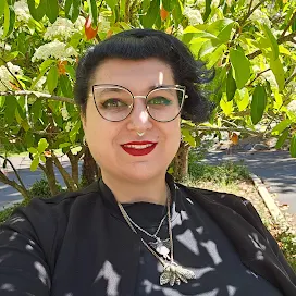 Lizzie Parkhurst - A Femme with short dark hair and glasses wears all black against a brightly light tree