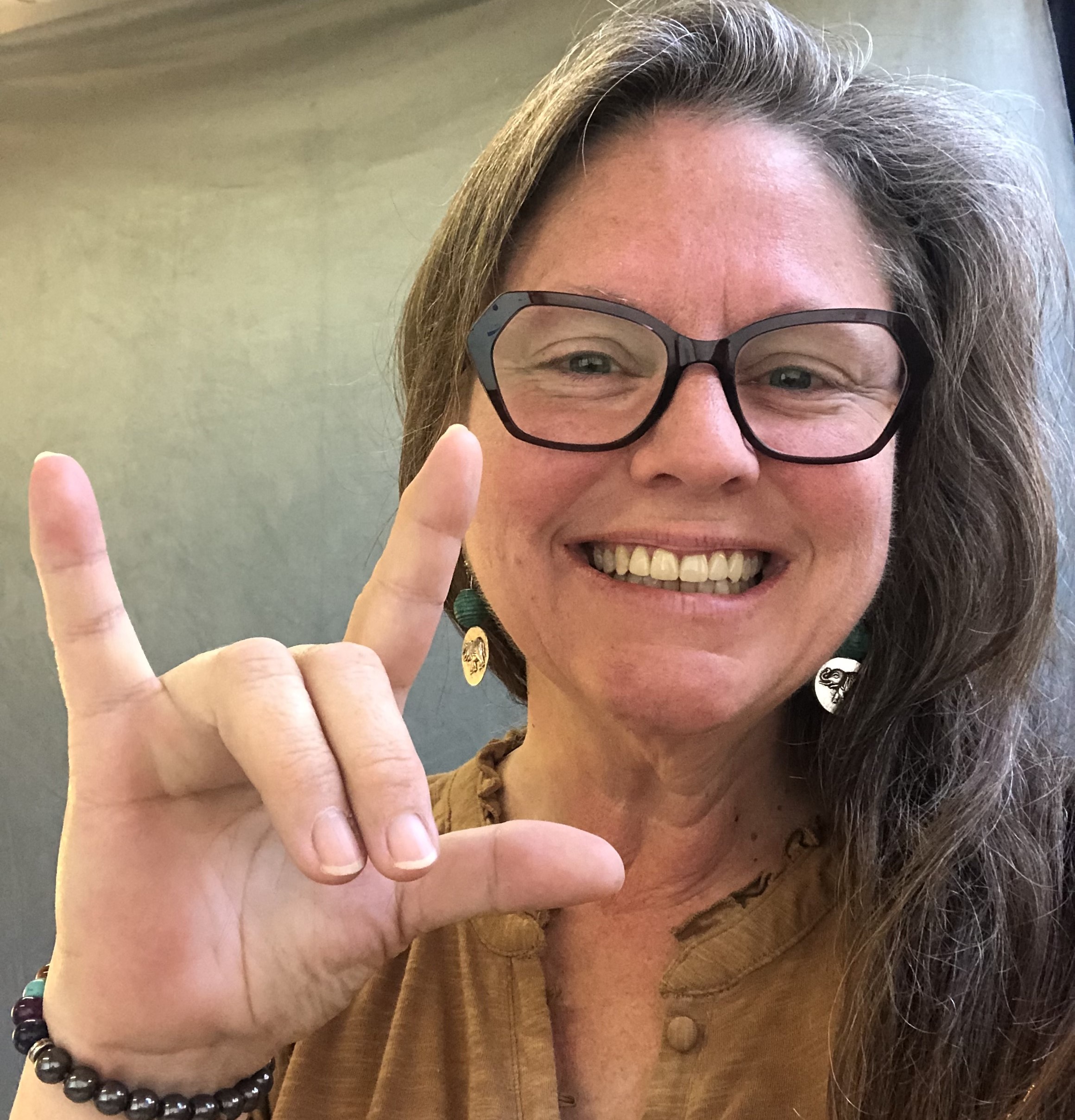 A Woman wearing glasses with long hair holds up the I love you sign in ASL
